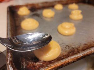 choux dough piped onto sheet pan with spoon flattening out top of one bun.